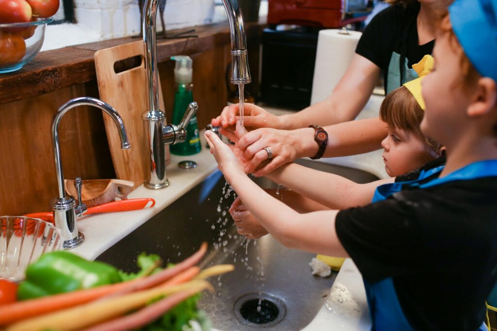 Mother helping children wash hands in kitchen sink.
